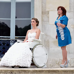 Femme en fauteuil roulant, portant une robe de mariée, à coté d’une autre femme, debout.