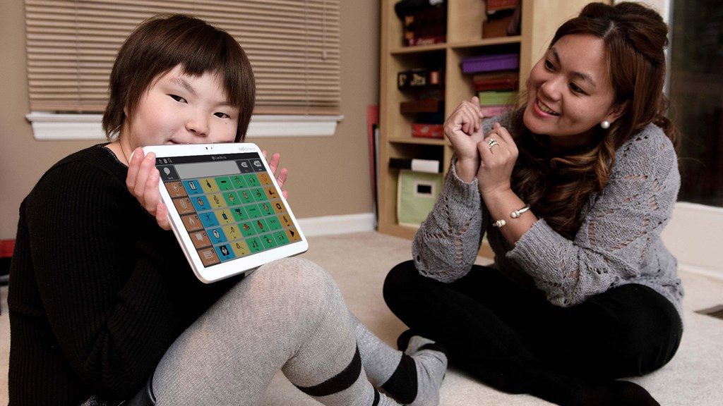 Diapo 3 : Gauche de l'image: un enfant tient une tablette Indi, affichant  le clavier de pictogrammes de l'application Core First. Droite de l'image: un adulte l'encourage.