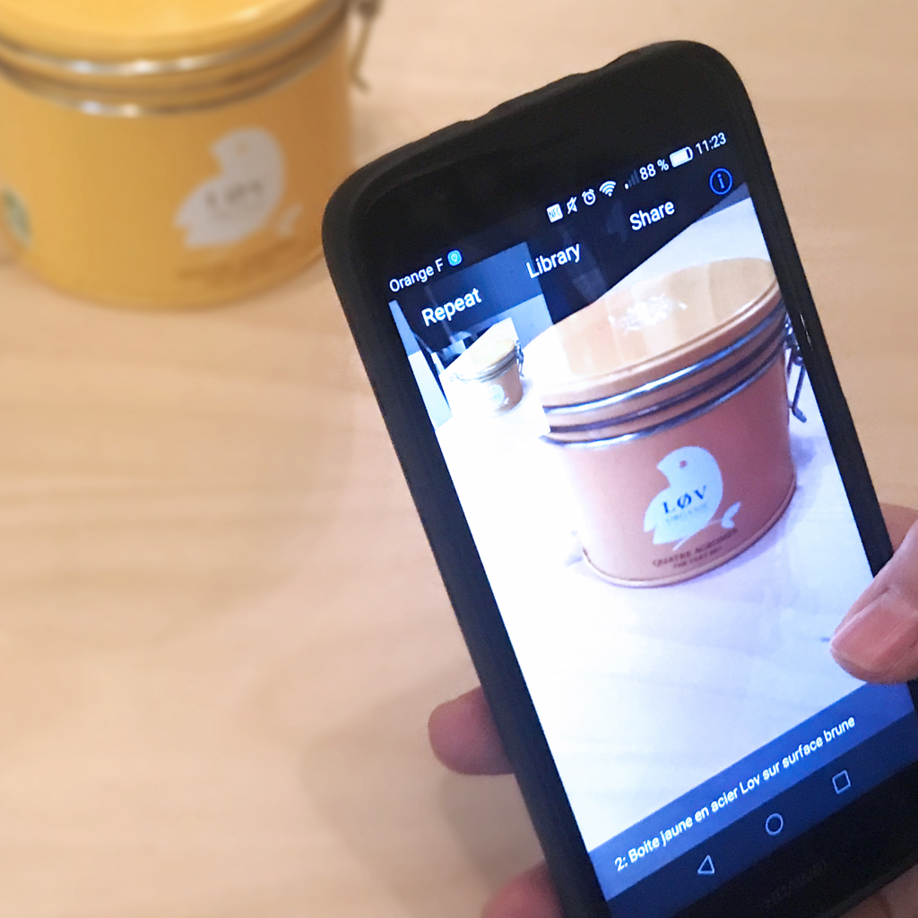 Diapo 2 : Personne utilisant l'application TapTapSee sur son smartphone pour identifier un objet posé sur une table. L'application indique 'Boite jaune en acier Lov sur surface brune'.