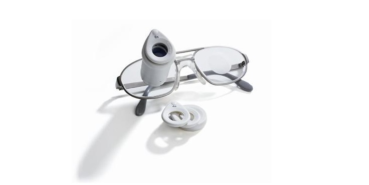 Diapo 3 : Lunettes ZEISS G2 BIOPTICS posées sur une surface blanche. Des lentilles de rechange pour la loupe intégrée sont posées sur le coté.