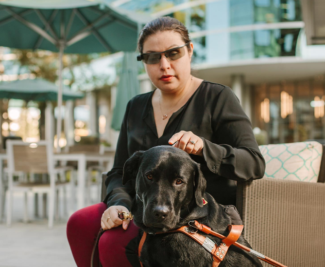 Diapo 3 : Personne équipée du dispositif Aira (lunettes et oreillettes) , assise, avec son chien guide d'aveugle.