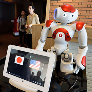 Robot Nao, sur une table à coté d’un ordinateur affichant les drapeau Américains et Japonais.