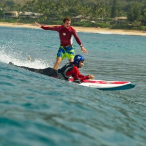 Homme handicapé des jambes qui fait du surf couché sur le ventre, avec derrière un autre homme qui surfe debout